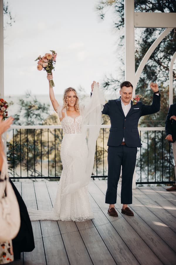 Bride Krystal and groom Brandon stand on the ceremony stage at Sandstone Point Hotel — Pavilion, holding hands raised in celebration. Krystal holds a bouquet of flowers in her other hand.