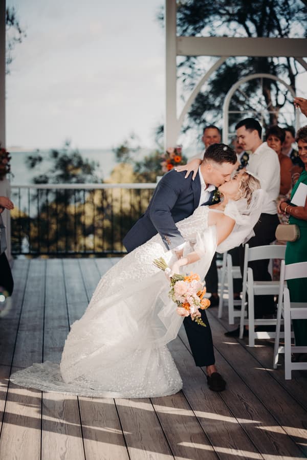 Bride Krystal and groom Brandon share a kiss on the ceremony stage at Sandstone Point Hotel — Pavilion, with seated guests watching.