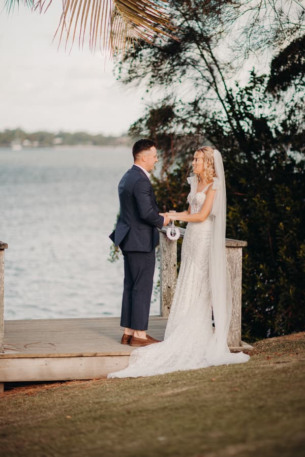 The bride Krystal and groom Brandon stand facing each other holding hands on a wooden platform near the water at Sandstone Point Hotel, with trees and foliage in the background.