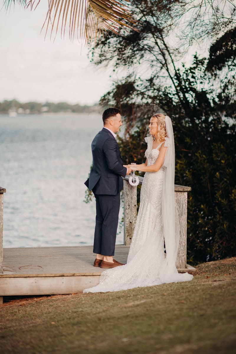 The bride Krystal and groom Brandon stand facing each other holding hands on a wooden platform near the water, with trees and foliage in the background. Cheap photo booth hire Brisbane by She Said Yes.
