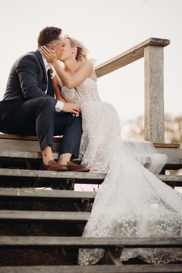 Bride Krystal and groom Brandon sit on wooden steps at Sandstone Point Hotel, sharing a kiss. Krystal wears a detailed white wedding gown with a long train, and Brandon is dressed in a dark suit with brown shoes.