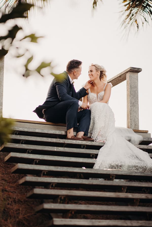 Bride Krystal and groom Brandon sit on wooden steps at Sandstone Point Hotel during their couple portraits session, with Krystal in a white wedding gown and Brandon in a dark suit.