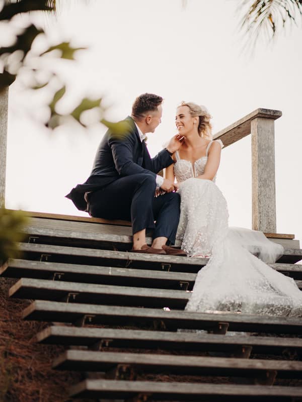 Bride Krystal and groom Brandon sit on wooden steps at Sandstone Point Hotel during their couple portraits session, with Krystal in a white wedding gown and Brandon in a dark suit.
