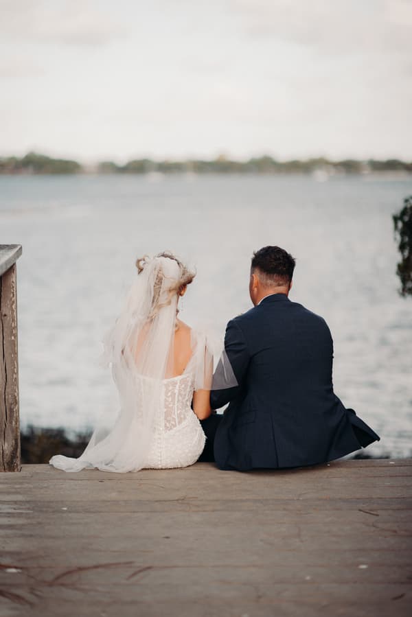 The bride and groom sit side by side on a wooden deck overlooking the water at Sandstone Point Hotel, viewed from behind.