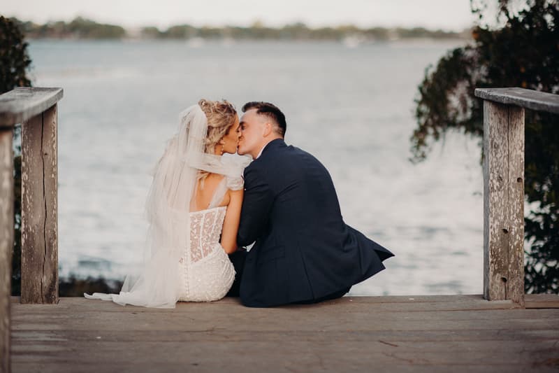 Bride Krystal and groom Brandon sit on a wooden dock at Sandstone Point Hotel, sharing a kiss with a body of water in the background.