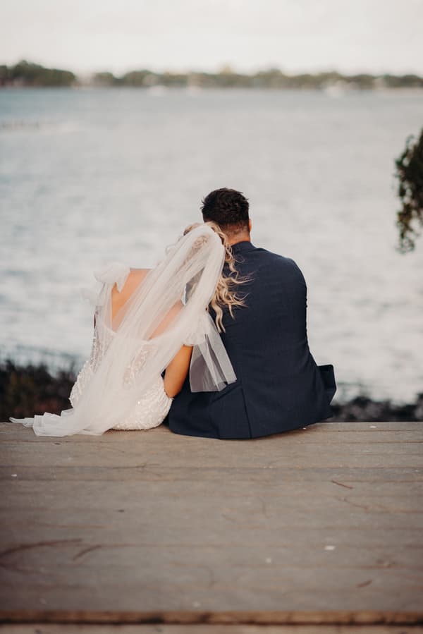 The bride and groom sit side by side on a wooden dock at Sandstone Point Hotel, facing a body of water. The bride wears a white dress with a veil, and the groom wears a dark suit.