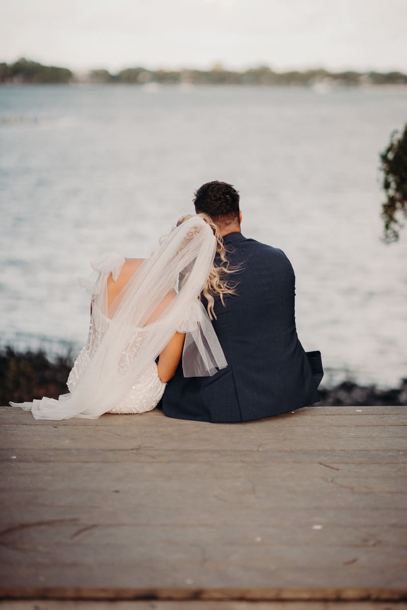 The bride and groom sit side by side on a wooden dock, facing a body of water. The bride wears a white dress with a veil, and the groom wears a dark suit. Sunset wedding portraits Brisbane by She Said Yes.