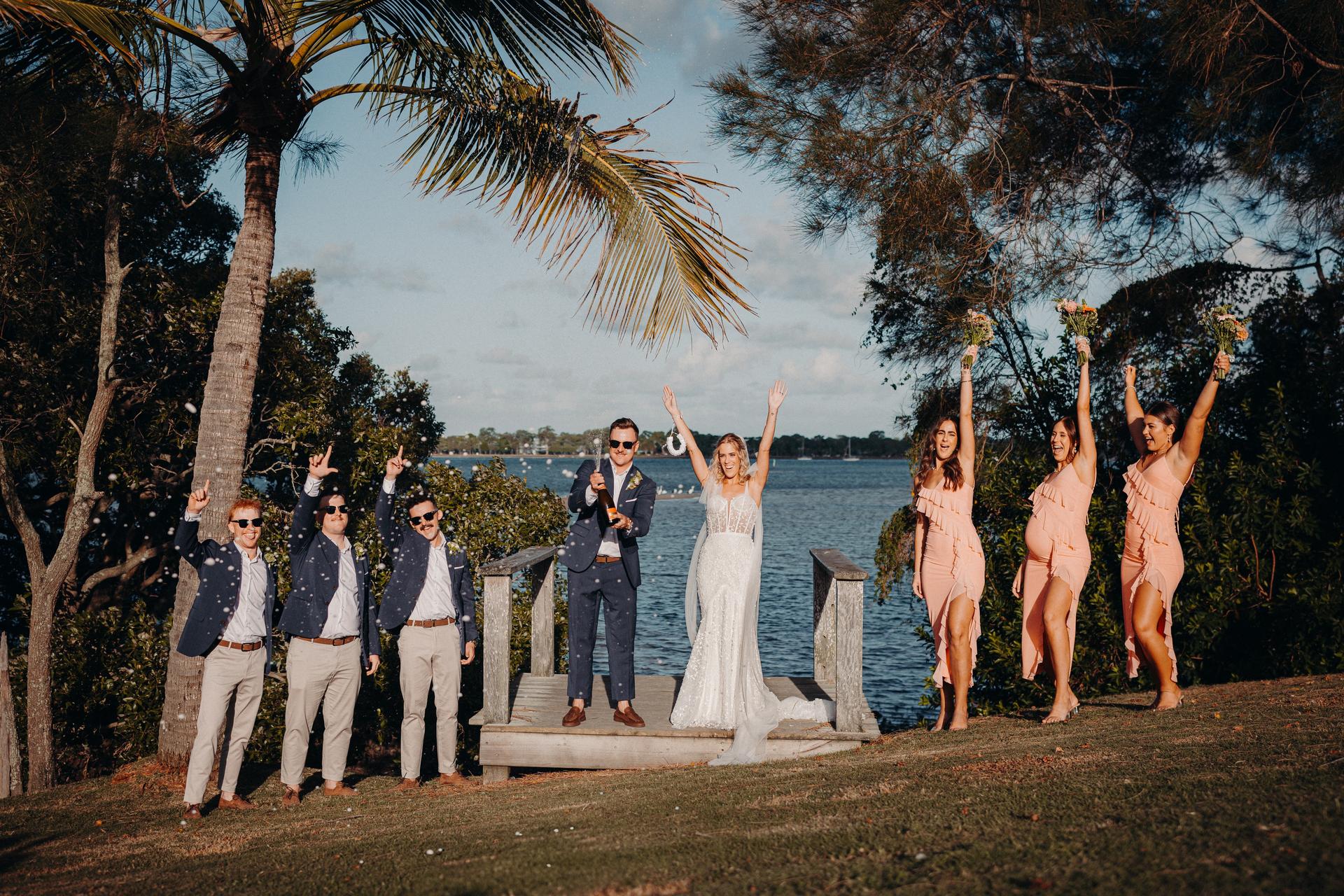 Krystal the bride and Brandon the groom stand on a wooden platform at Sandstone Point Hotel by the water, surrounded by three groomsmen in navy blazers and beige pants on the left and three bridesmaids in peach dresses holding bouquets on the right. Brandon is popping a bottle of champagne while Krystal and the bridesmaids raise their arms in celebration.