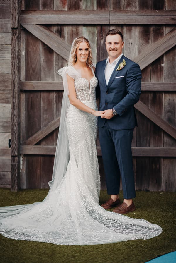 The bride Krystal in a white lace wedding gown and veil stands holding hands with the groom Brandon in a navy blue suit and brown shoes in front of a large wooden barn door at Sandstone Point Hotel.
