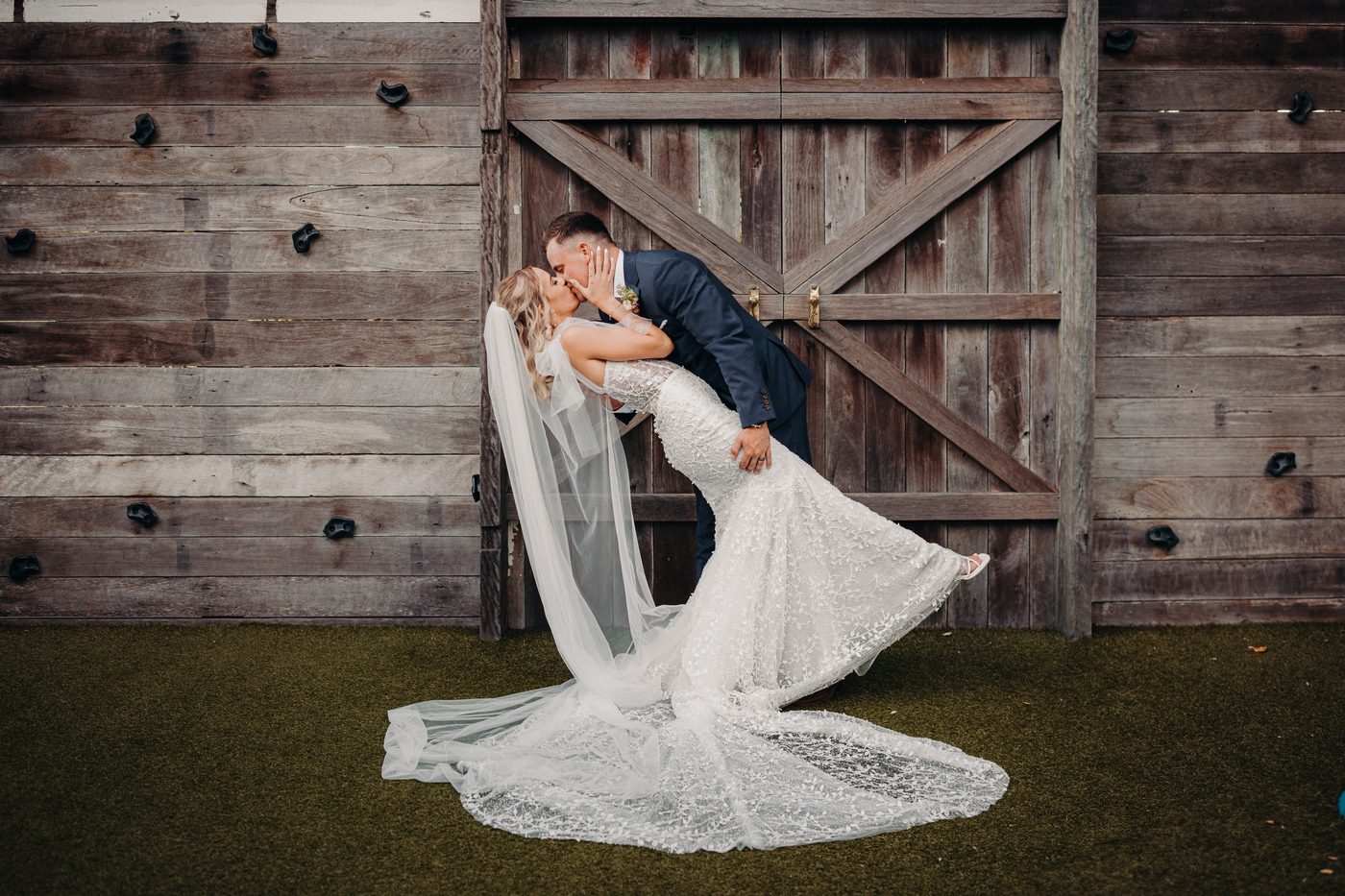 The bride Krystal and groom Brandon share a kiss in front of a large rustic wooden door at Sandstone Point Hotel during their couple portraits session. Krystal is wearing a lace wedding gown with a long veil, and Brandon is dressed in a dark suit.
