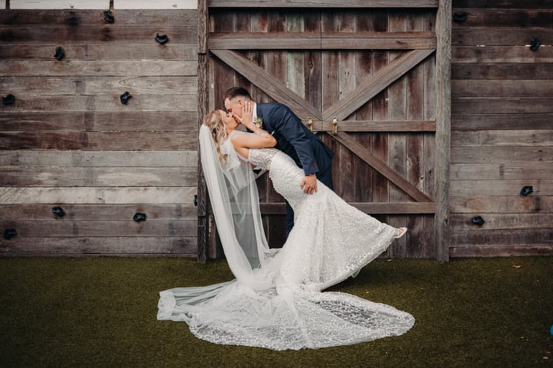 The bride Krystal and groom Brandon share a kiss in front of a large rustic wooden door at Sandstone Point Hotel during their couple portraits session. Krystal is wearing a lace wedding gown with a long veil, and Brandon is dressed in a dark suit.