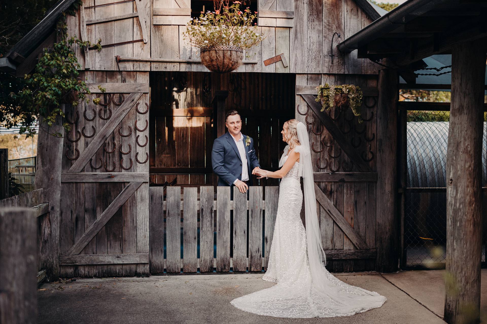 Bride Krystal and groom Brandon stand holding hands in front of a rustic wooden barn door decorated with horseshoes and hanging plants at Sandstone Point Hotel.