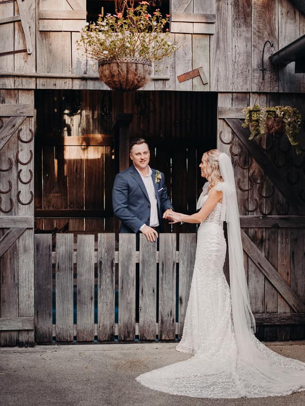 Bride Krystal and groom Brandon stand holding hands in front of a rustic wooden barn door decorated with horseshoes and hanging plants at Sandstone Point Hotel.