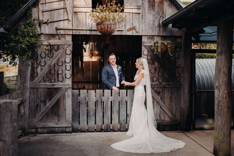 Bride Krystal and groom Brandon stand holding hands in front of a rustic wooden barn door decorated with horseshoes and hanging plants at Sandstone Point Hotel.