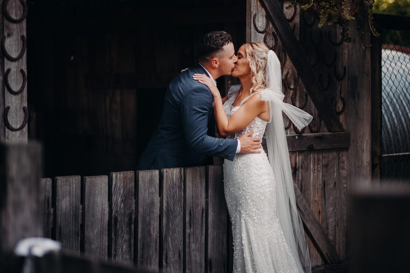 Bride Krystal and groom Brandon share a kiss in front of a rustic wooden gate. Popular wedding seasons 2026 by She Said Yes.