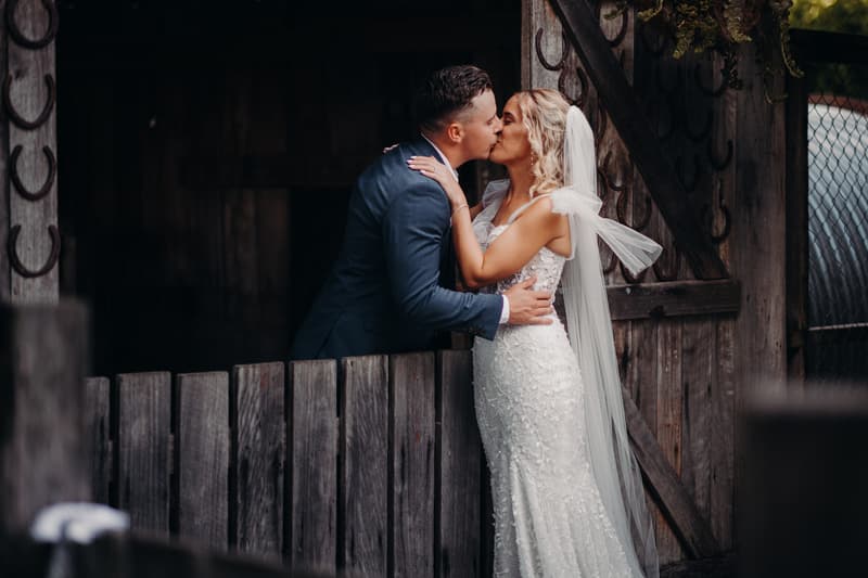 Bride Krystal and groom Brandon share a kiss in front of a rustic wooden gate at Sandstone Point Hotel during their couple portraits session.