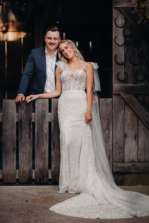 Krystal in a white lace wedding gown with a long train and Brandon in a blue suit pose together leaning on a rustic wooden fence at Sandstone Point Hotel.