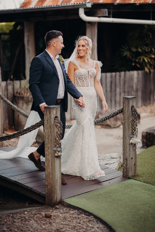 Bride Krystal and groom Brandon hold hands and walk across a small wooden bridge at Sandstone Point Hotel, with Krystal wearing a lace wedding gown and Brandon in a dark suit.