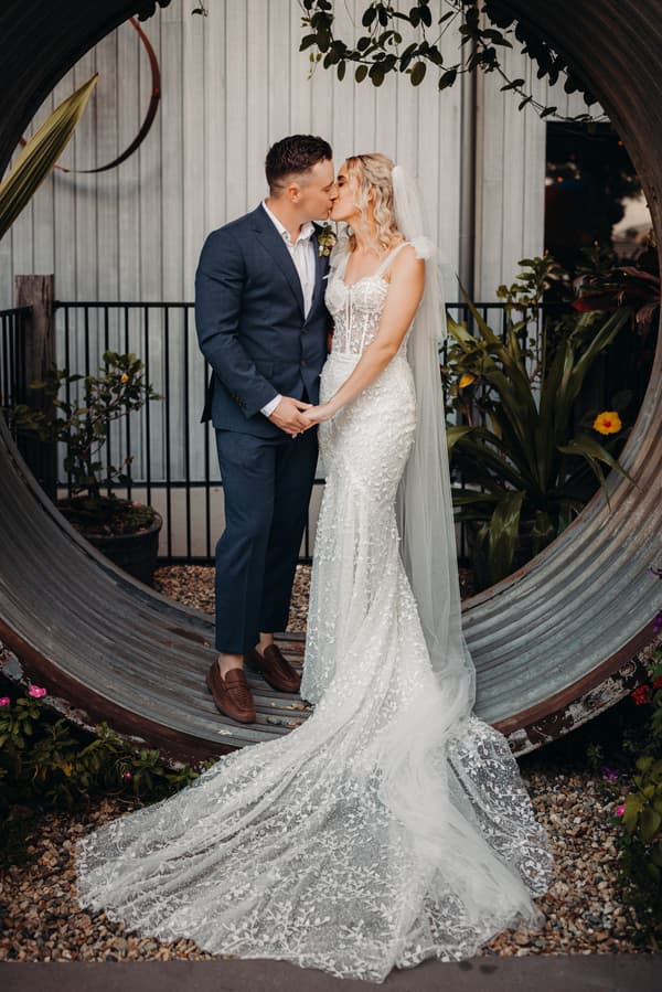 Bride Krystal and groom Brandon kiss while holding hands, standing inside a large circular metal structure at Sandstone Point Hotel. Krystal wears a white lace wedding gown with a long train and veil, and Brandon wears a dark blue suit with brown shoes.