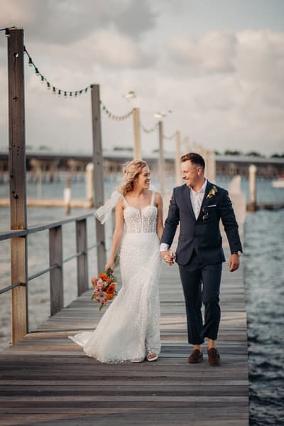 The bride Krystal in a white wedding gown holding a bouquet and the groom Brandon in a navy suit walk hand in hand along a wooden pier at Sandstone Point Hotel, with water and posts with string lights in the background.