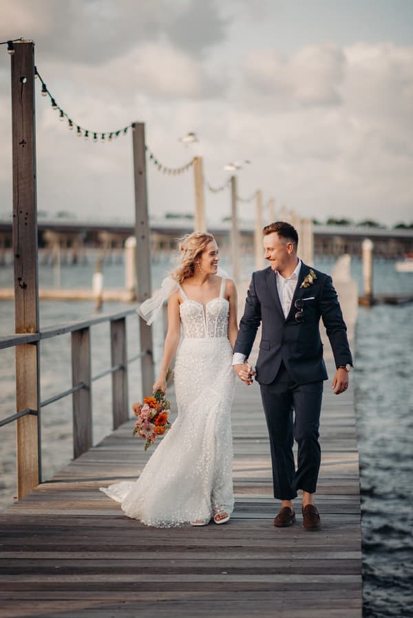 The bride Krystal in a white wedding gown holding a bouquet and the groom Brandon in a navy suit walk hand in hand along a wooden pier at Sandstone Point Hotel, with water and posts with string lights in the background.