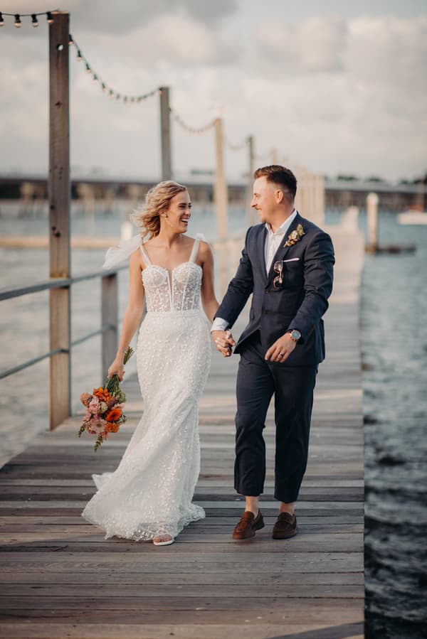 The bride Krystal in a white lace wedding gown holding a bouquet and the groom Brandon in a navy suit walk hand in hand along a wooden pier at Sandstone Point Hotel.