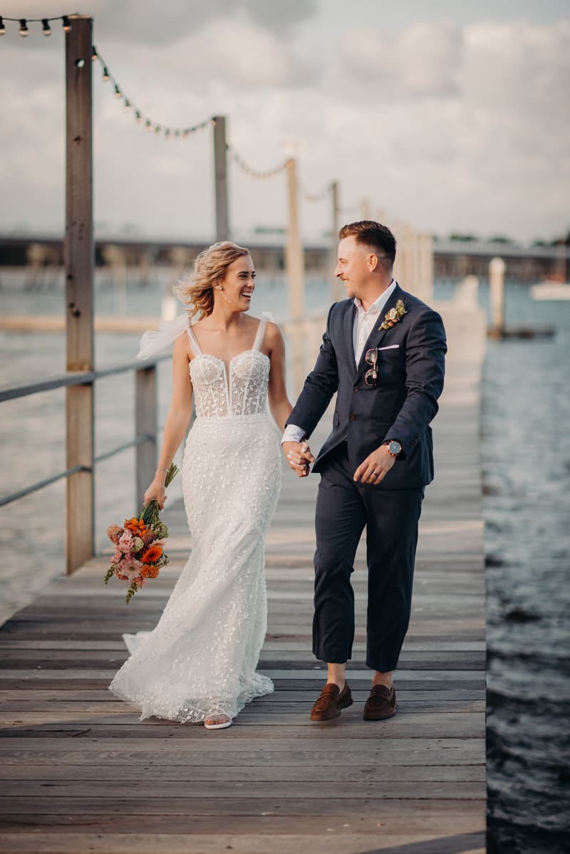 The bride Krystal in a white lace wedding gown holding a bouquet and the groom Brandon in a navy suit walk hand in hand along a wooden pier. Shortest day wedding timeline by She Said Yes.