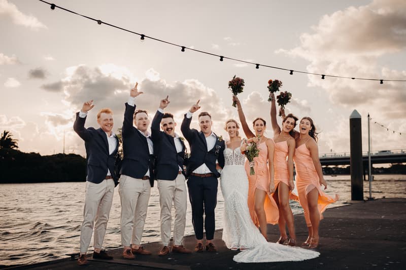 Krystal the bride and Brandon the groom pose with their bridal party at Sandstone Point Hotel near the water during sunset. The groom and groomsmen wear navy jackets and light pants, while the bride and bridesmaids wear white and peach dresses respectively, with the bridesmaids holding bouquets raised in the air.