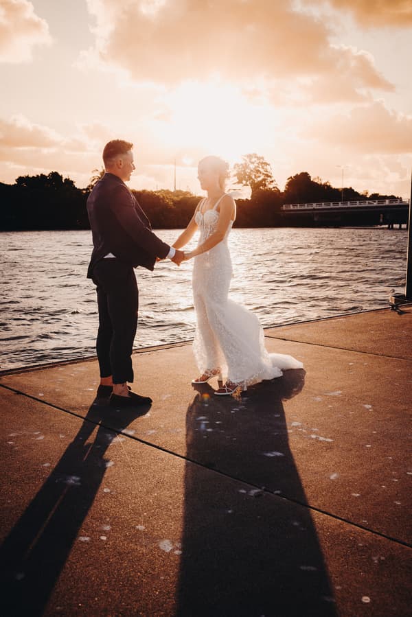 Bride Krystal and groom Brandon hold hands facing each other on a pier at Sandstone Point Hotel during sunset, with water and trees in the background.
