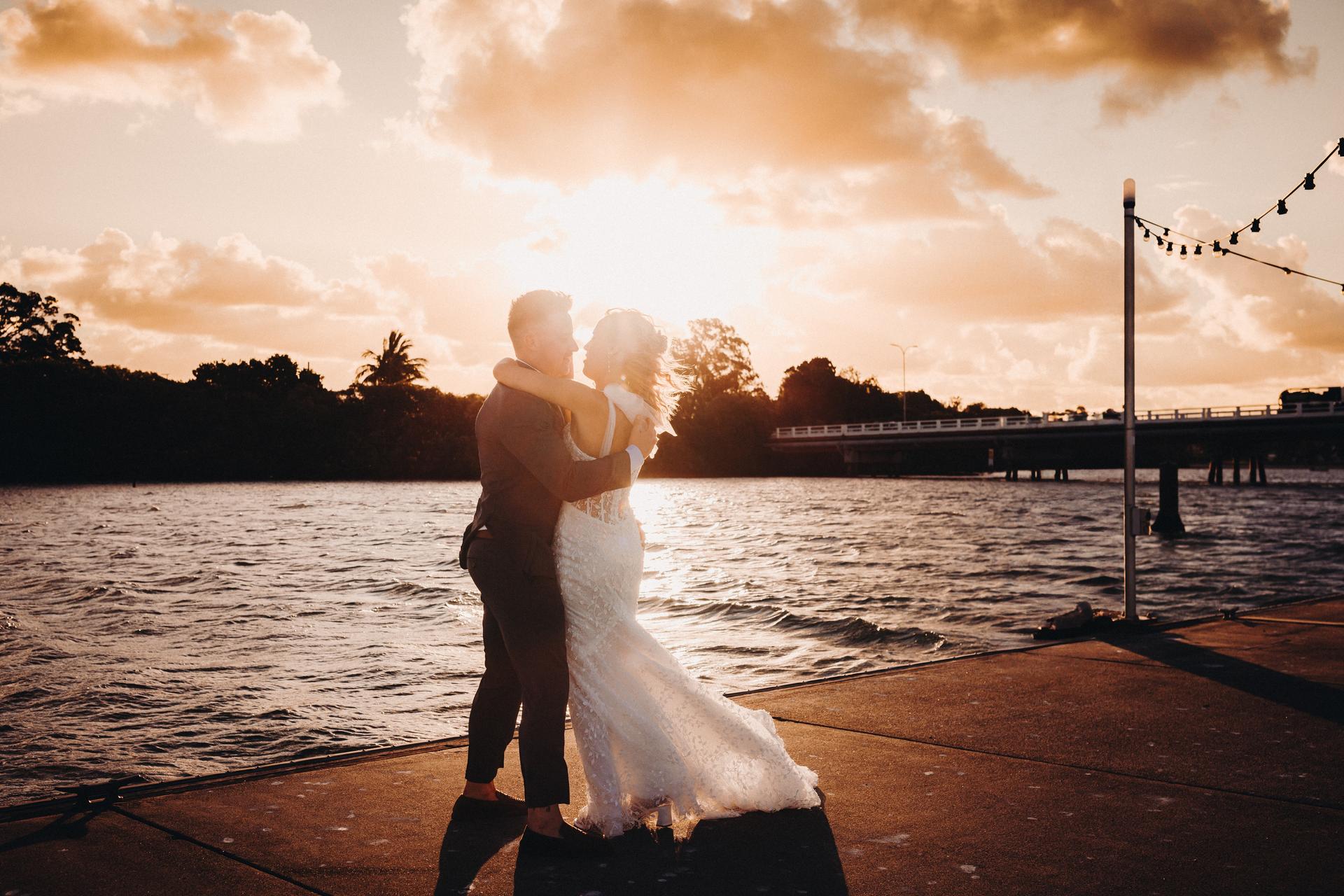 Bride Krystal and groom Brandon embrace on a waterfront pier at Sandstone Point Hotel during sunset with a bridge and trees in the background.