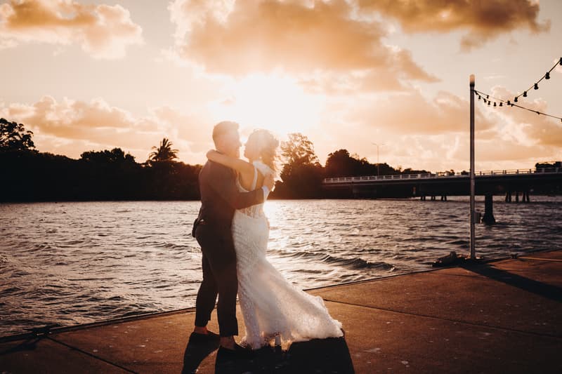 Bride Krystal and groom Brandon embrace on a waterfront pier at Sandstone Point Hotel during sunset with a bridge and trees in the background.
