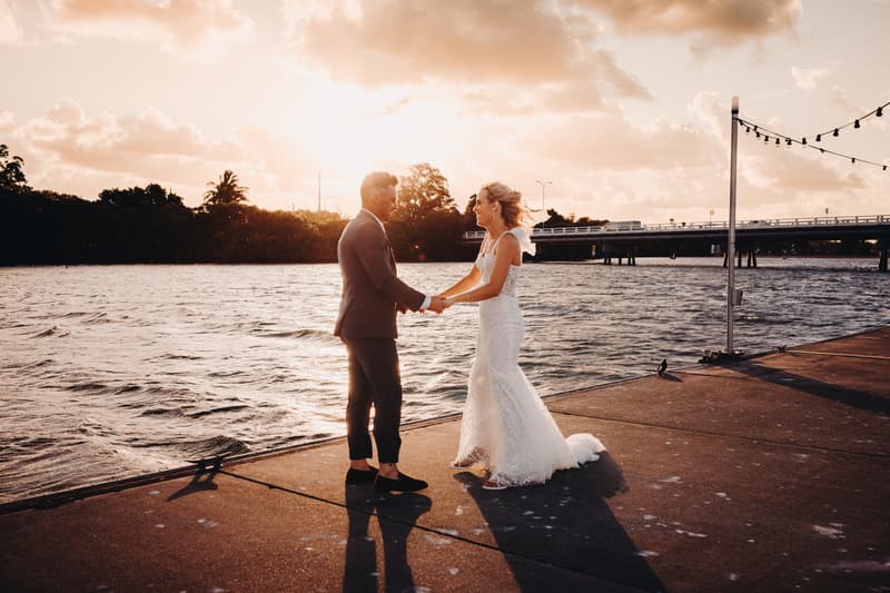 Bride Krystal and groom Brandon hold hands facing each other on a waterfront pier at Sandstone Point Hotel during sunset.