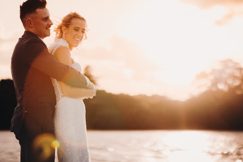 The bride Krystal and groom Brandon embrace during a couple portrait session at Sandstone Point Hotel with a sunset over water in the background.