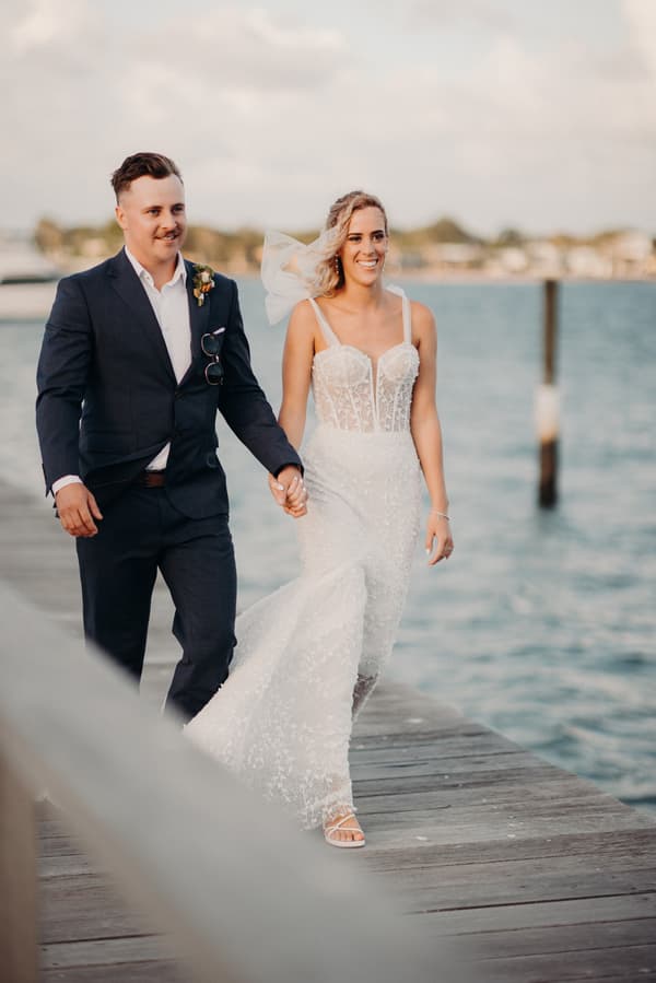 Bride Krystal and groom Brandon walk hand in hand on a wooden pier at Sandstone Point Hotel, with water and distant shoreline in the background.