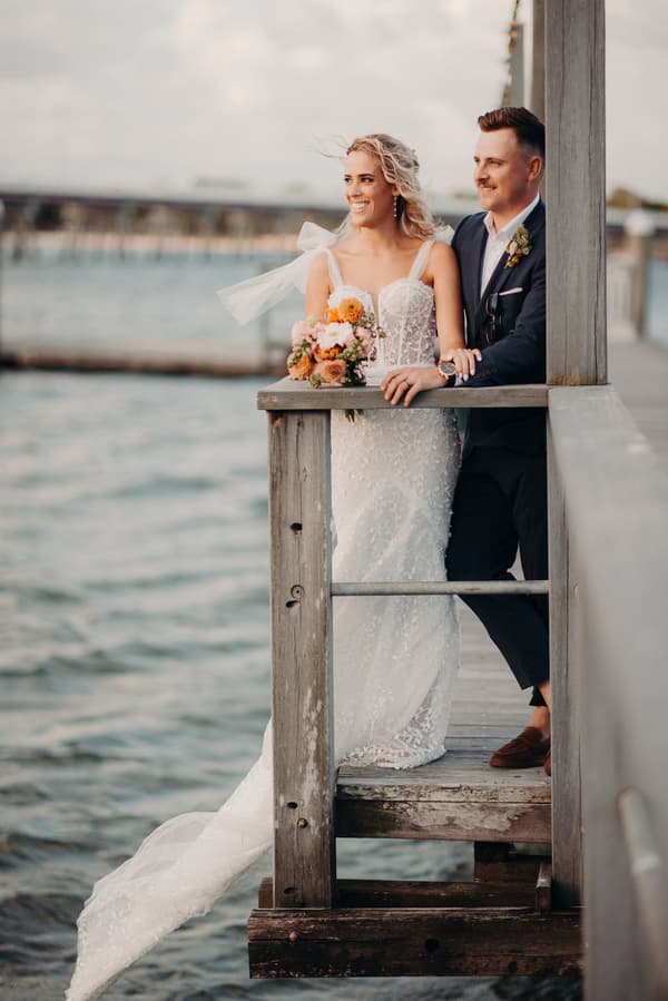 Bride Krystal in a white lace wedding gown holding a bouquet and groom Brandon in a dark suit lean on a wooden railing overlooking water at Sandstone Point Hotel.