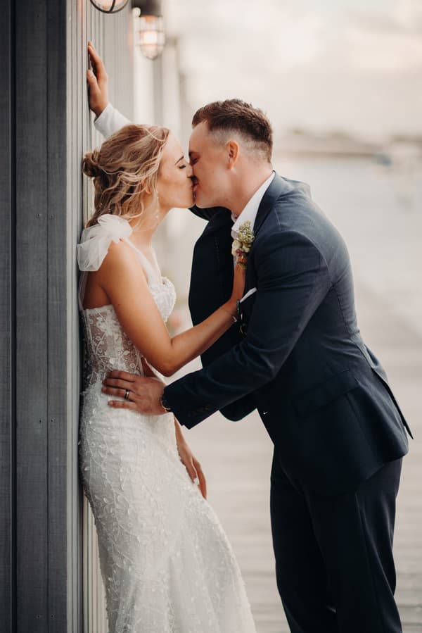 The bride Krystal and groom Brandon share a kiss during their couple portraits at Sandstone Point Hotel. Krystal is wearing a white lace wedding gown with sheer shoulder details, and Brandon is dressed in a dark suit with a boutonniere. They are positioned against a wooden wall with outdoor lighting fixtures visible.