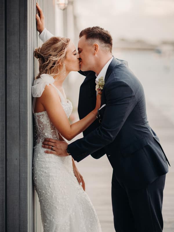 The bride Krystal and groom Brandon share a kiss during their couple portraits at Sandstone Point Hotel. Krystal is wearing a white lace wedding gown with sheer shoulder details, and Brandon is dressed in a dark suit with a boutonniere. They are positioned against a wooden wall with outdoor lighting fixtures visible.