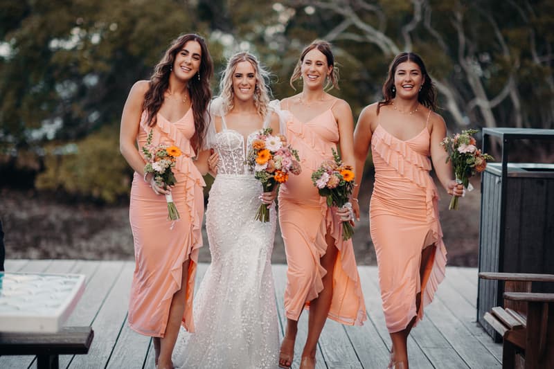 The bride Krystal stands with three bridesmaids in peach dresses holding bouquets at Sandstone Point Hotel outdoors on a wooden deck.