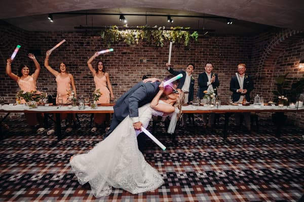 Bride Krystal and groom Brandon share a kiss in a dance dip at the Sandstone Point Hotel — Cellar reception stage, while bridesmaids and groomsmen stand behind a long table holding light sticks and applauding.