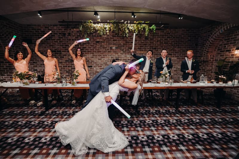 Bride Krystal and groom Brandon share a kiss in a dance dip at the Sandstone Point Hotel — Cellar reception stage, while bridesmaids and groomsmen stand behind a long table holding light sticks and applauding.