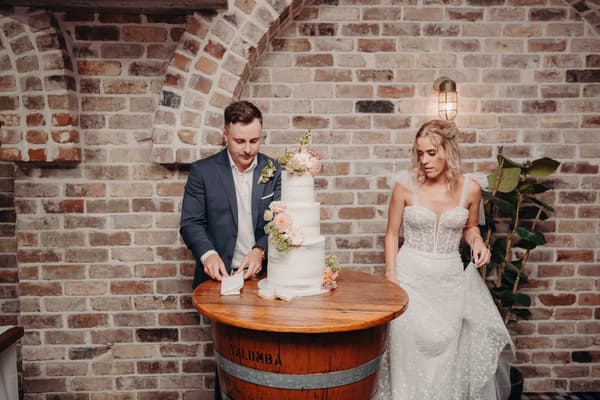 Bride Krystal and groom Brandon stand beside a three-tier wedding cake on a wooden barrel table at Sandstone Point Hotel — Cellar.