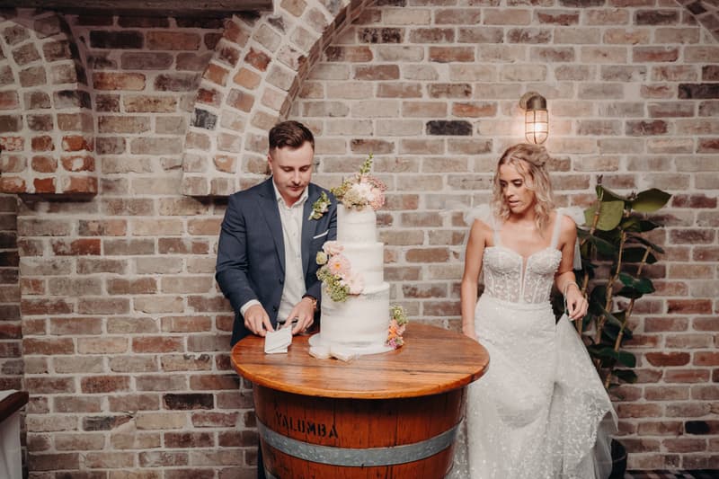 Bride Krystal and groom Brandon stand beside a three-tier wedding cake on a wooden barrel table at Sandstone Point Hotel — Cellar.