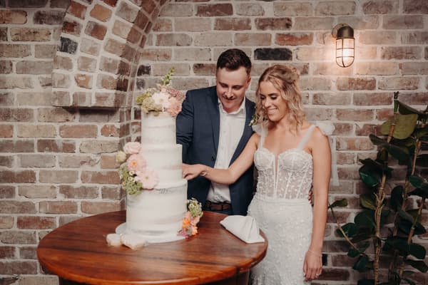 Bride Krystal and groom Brandon cut their wedding cake at Sandstone Point Hotel — Cellar, standing beside a round wooden table with a four-tiered white cake decorated with flowers, against a brick wall with a wall light and a plant nearby.