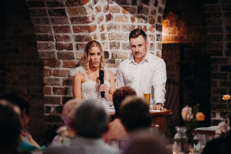 Bride Krystal and groom Brandon stand at a wooden podium against a brick wall at Sandstone Point Hotel — Cellar, with Krystal holding a microphone and Brandon standing beside her with a glass of beer, addressing seated guests.