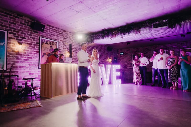 Bride Krystal and groom Brandon stand together on the reception stage at Sandstone Point Hotel — Cellar, with guests watching and a DJ at a booth nearby. Illuminated letters spelling LOVE are visible behind them.