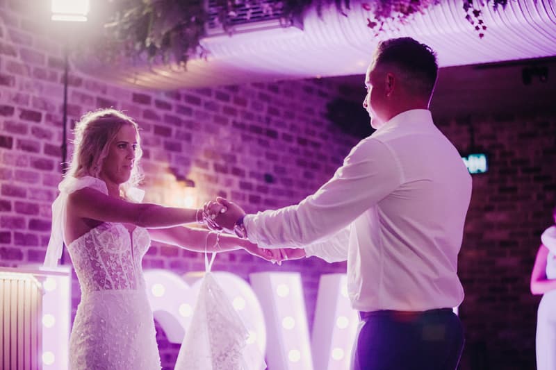 Bride Krystal and groom Brandon hold hands facing each other on the reception stage at Sandstone Point Hotel — Cellar, with illuminated letters in the background.