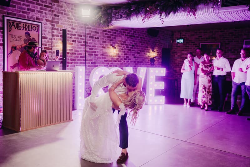 Bride Krystal and groom Brandon share a dance on the floor at Sandstone Point Hotel — Cellar with illuminated LOVE letters behind them and guests watching in the background near a DJ booth.