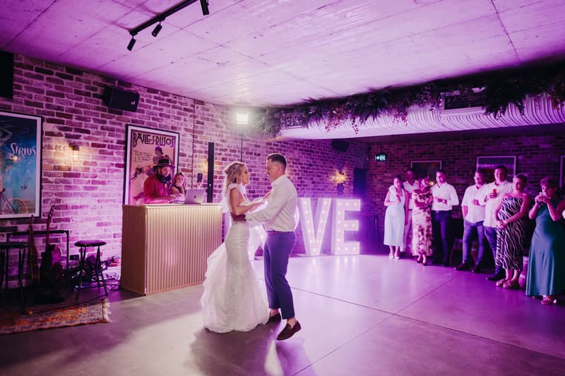 Bride Krystal and groom Brandon dance together on the reception stage at Sandstone Point Hotel — Cellar, with guests watching and a DJ at the booth near illuminated LOVE letters.