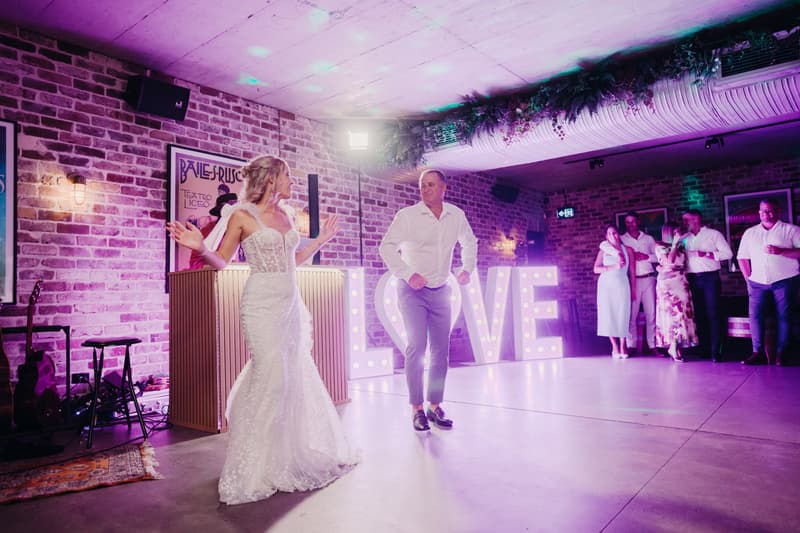 Bride Krystal and groom Brandon dance on the reception stage at Sandstone Point Hotel — Cellar with illuminated LOVE letters behind them and guests watching in the background.
