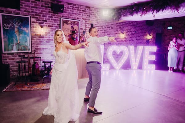 Bride Krystal and groom Brandon dance together on the reception stage at Sandstone Point Hotel — Cellar, with illuminated LOVE letters in the background and guests watching.